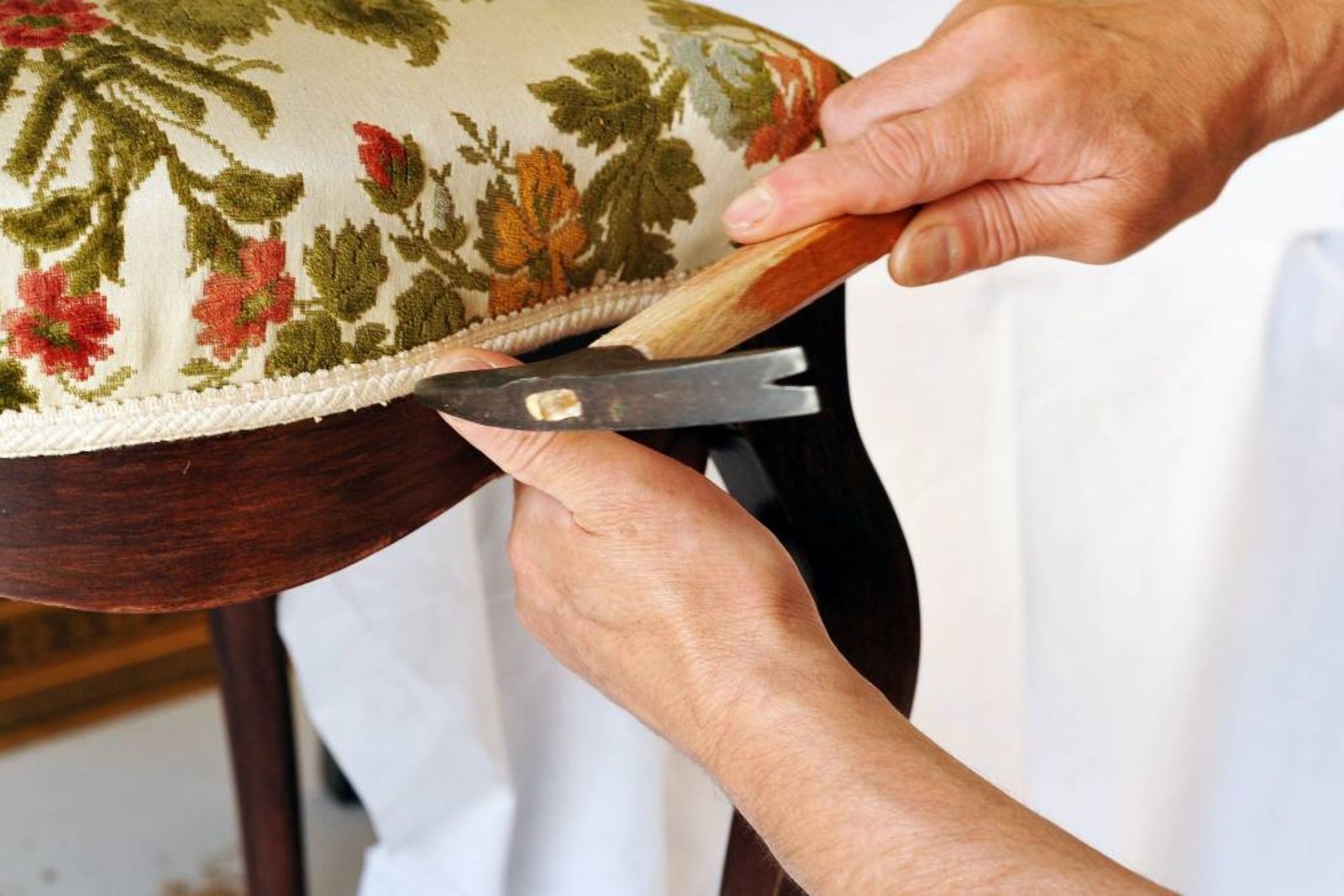 A person carefully reupholstering a chair, using a tool to trim fabric with floral patterns on the seat.