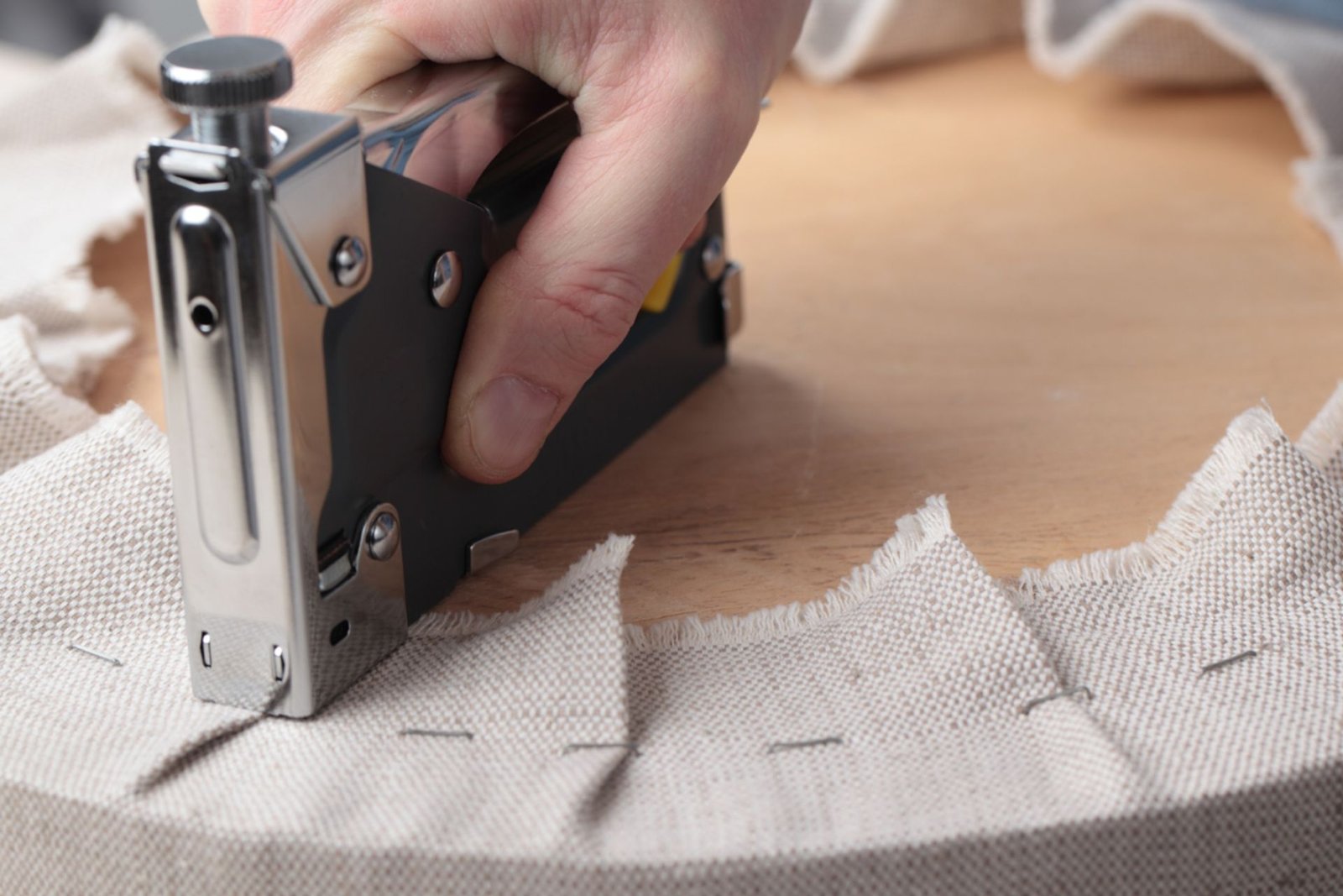 A hand using a staple gun to attach fabric around a wooden frame, showcasing the crafting process of upholstery
