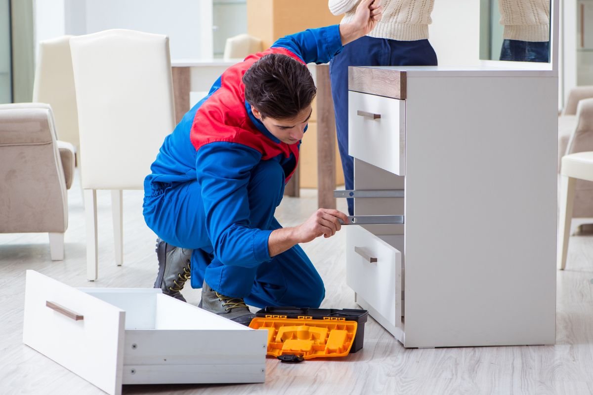 A man in blue overalls repairs a cabinet, showcasing his skills in furniture service.