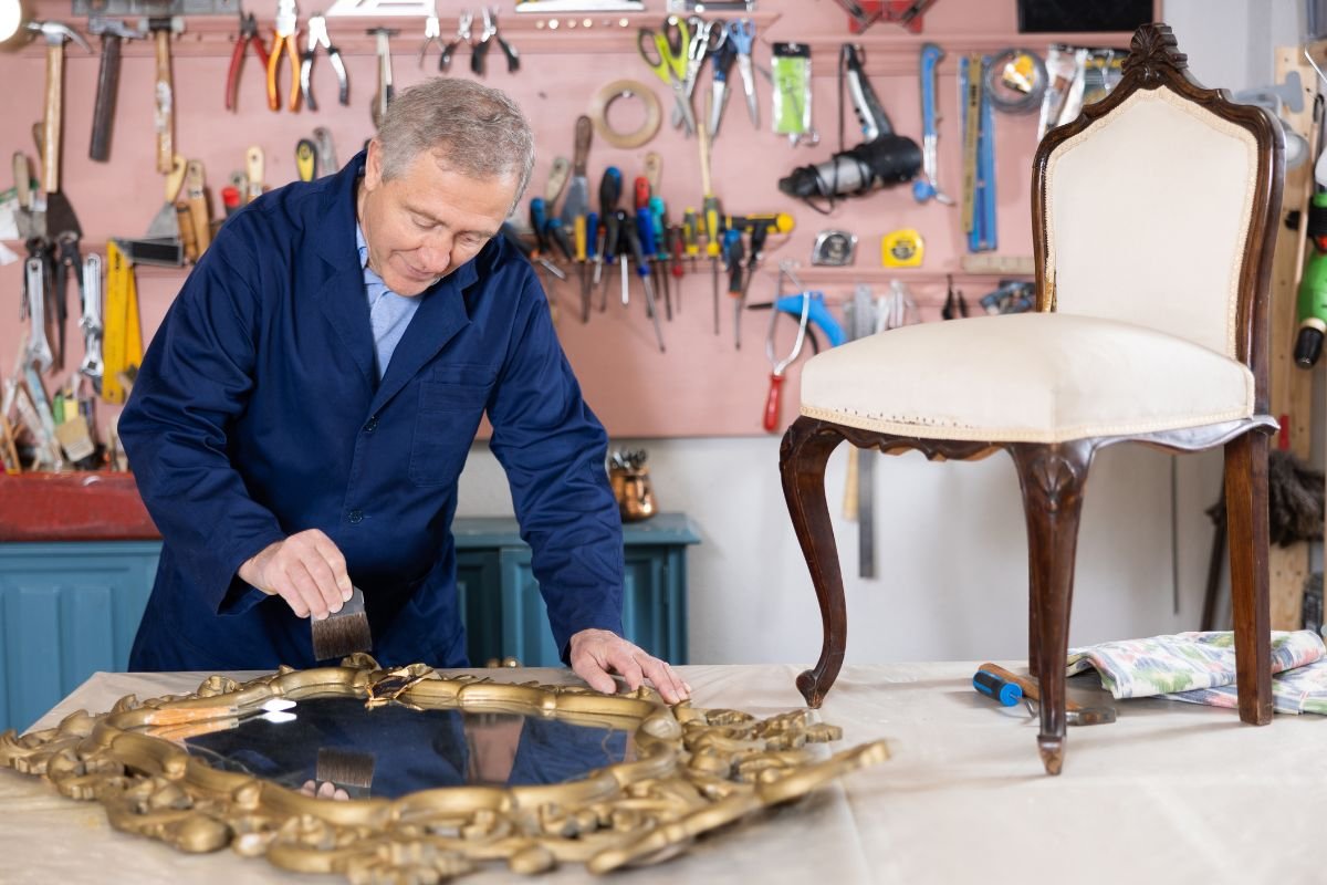 A man repairs a chair in a workshop, surrounded by tools and materials, showcasing furniture service craftsmanship.
